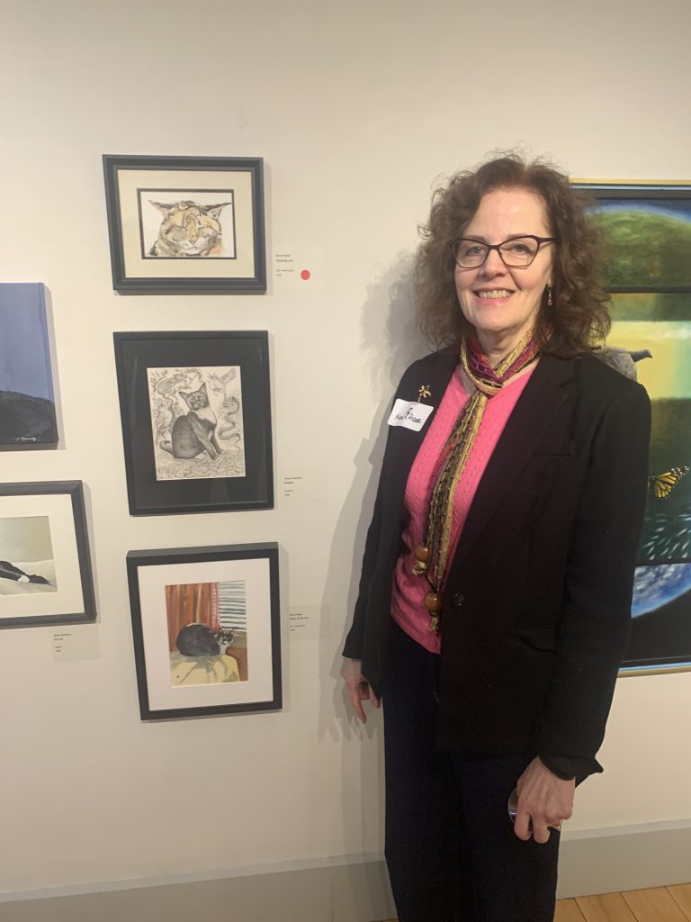 Karen R. Stands beside a display of paintings of cats, 2 of which were created by her